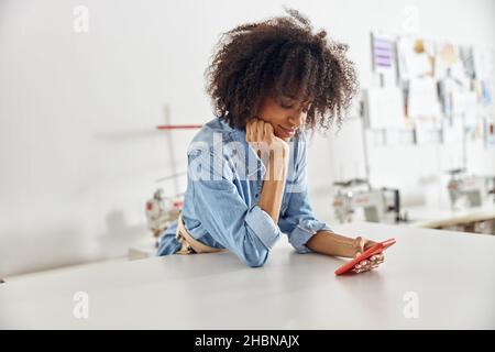 Le joli designer afro-américain en chemise denim utilise un téléphone portable dans un atelier de couture spacieux Banque D'Images