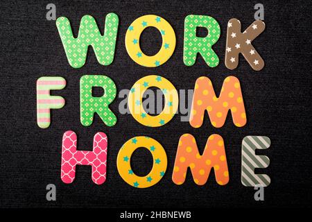 Carte avec texte « Work from Home » composée de lettres en bois aux couleurs vives et mélangées sur un tissu noir foncé texturé pouvant servir de message Banque D'Images