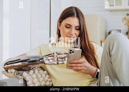 Une femme heureuse avec un bras prothétique bionique est assise sur le canapé avec un téléphone et communique en ligne avec des amis ou surfent sur Internet ou effectue des achats en ligne dans un T-shirt jaune sur le fond d'une cuisine lumineuse Banque D'Images