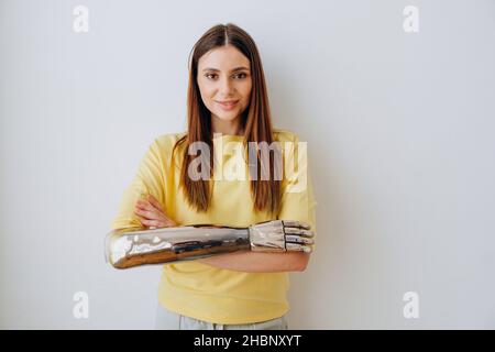 Jolie jeune femme blonde souriante avec un élégant bras de prothèse bionique regarde dans l'appareil photo debout sur fond gris clair gros plan Banque D'Images