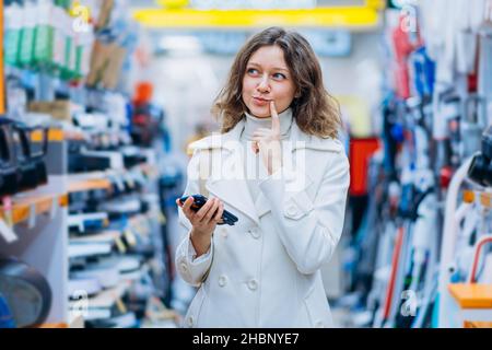 belle femme shopper dans un manteau blanc a pensé avec un téléphone dans la main dans un magasin d'appareils à la maison dans un supermarché, l'émotion de la joie et la difficulté de choisir avant d'acheter Banque D'Images