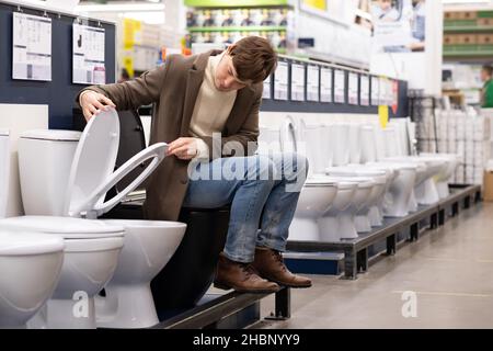 L'homme européen en manteau brun choisit un bol de toilette en céramique dans un supermarché de construction, assis sur les toilettes Banque D'Images