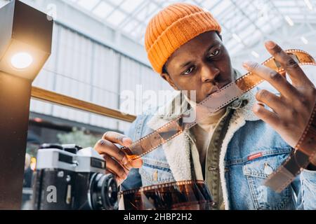 Photographe afro-américain concentré en veste denim vérifie le film vintage à la table se concentre sur l'ancien appareil photo vue rapprochée Banque D'Images