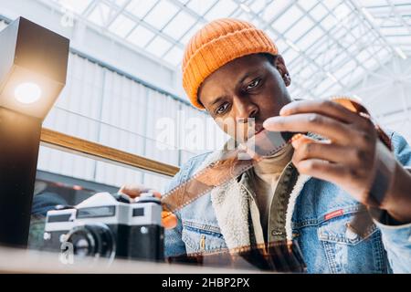 Photographe afro-américain concentré en veste denim vérifie le film vintage à la table se concentre sur l'ancien appareil photo vue rapprochée Banque D'Images