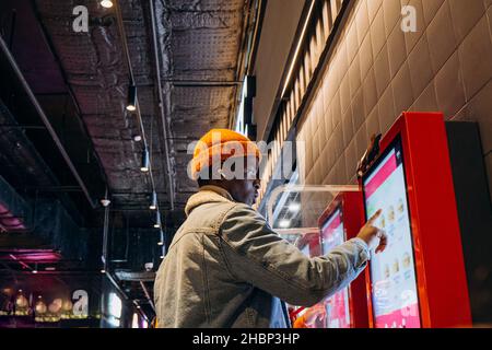 Un homme afro-américain souriant dans une veste en denim chaud avec écouteurs sans fil utilise un kiosque en libre-service pour commander des en-cas au café Banque D'Images