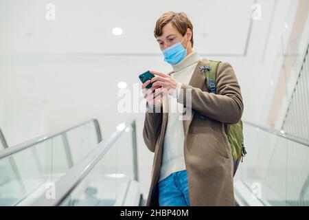 un homme moderne, portant un manteau marron avec un sac à dos et un masque, fait un escalier mécanique dans un centre commercial et regarde le téléphone entre ses mains Banque D'Images