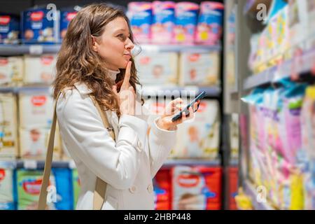 Belle femme caucasienne réussie dans un manteau blanc choisit des couches dans un supermarché avec un téléphone, en méditer une liste d'achats Banque D'Images