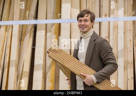 portrait d'un homme réussi dans un manteau brun dans un magasin de matériel regardant la caméra avec des planches en bois dans ses mains Banque D'Images