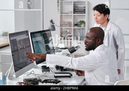 Un jeune scientifique confiant pointe sur un schéma de micro-puce agrandi sur un écran d'ordinateur au cours d'une discussion avec une collègue féminine Banque D'Images