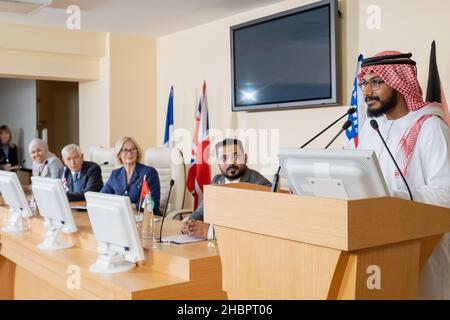 Jeune homme arabe confiant dans des vêtements traditionnels debout par tribune et parlant en micro lors d'une conférence politique Banque D'Images