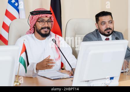 Jeune délégué arabe confiant parlant en micro lors d'une conférence ou d'un sommet politique contre un collègue étranger Banque D'Images