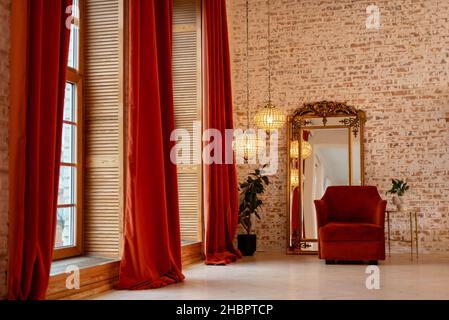 Un intérieur français moderne avec des rideaux en velours côtelé bordeaux, un fauteuil, un miroir doré vintage et des lustres en cristal contre un mur de briques.Donc Banque D'Images