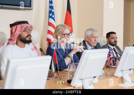 Une femme blonde mûre, qui fait un discours pour le public tout en étant assise à table entre plusieurs collègues étrangers Banque D'Images