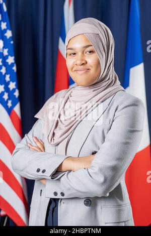 Jeune femme musulmane bien habillée en hijab et élégante costume debout devant la caméra sur fond de plusieurs drapeaux Banque D'Images