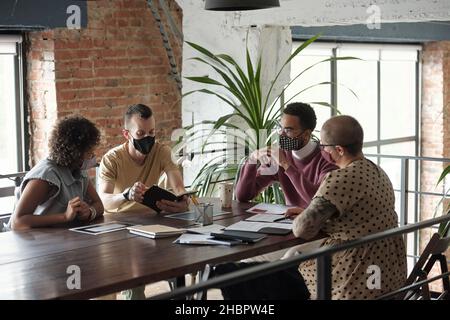 Groupe de jeunes collègues dans les masques de protection planifier le travail et discuter de nouvelles idées lors de la réunion tout en étant assis à table Banque D'Images