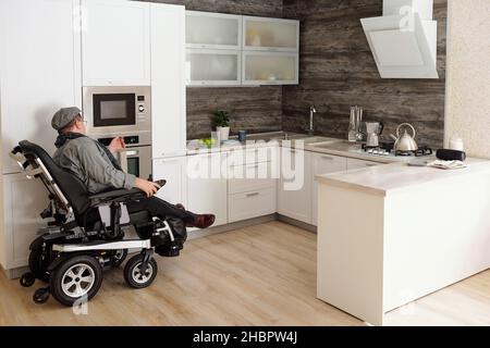 Homme d'âge moyen avec un handicap assis au four électrique dans une grande cuisine confortable dans un environnement domestique Banque D'Images