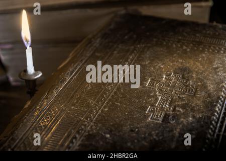 Une bougie dans un chandelier illumine la couverture d'une bible antique avec une croix en relief et un livre dans le fond à l'ombre Banque D'Images