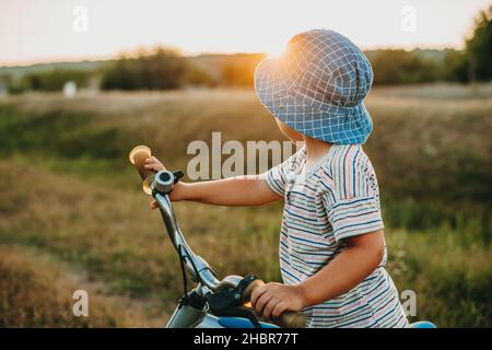 Vue d'un petit garçon prenant ses deux mains sur le vélo avec sa tête tournée portant un chapeau bleu.Un mode de vie sain.Journée des enfants. Banque D'Images