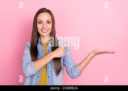 Photo de brunette optimiste femme mûre tenue indiquent l'espace vide porter Jean veste isolée sur fond rose pastel Banque D'Images