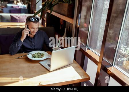 homme d'affaires parle au téléphone, tout en déjeunant et en travaillant sur un ordinateur portable dans le restaurant. Banque D'Images