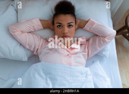 Femme afro-américaine fatiguée souffrant de dépression et de stress, allongé sur un lit blanc à la maison, vue de dessus Banque D'Images