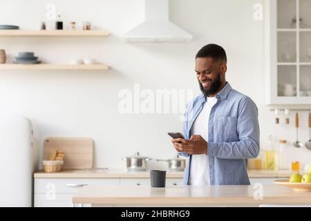 Un jeune homme afro-américain souriant et barbu dans un intérieur minimaliste de cuisine lit l'actualité Banque D'Images