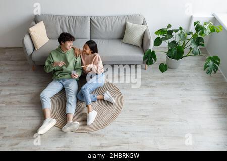 Vue de dessus de jeune asiatique souriant homme et femme regardant l'écran de smartphone, assis sur le sol à la maison, espace de copie Banque D'Images