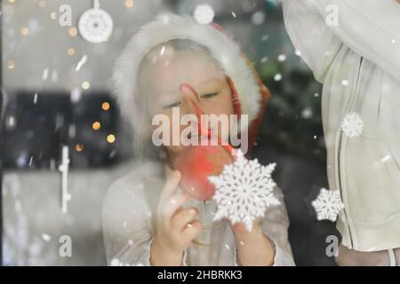 Une fille portant un chapeau de père noël regarde les flocons de neige sur une fenêtre Banque D'Images