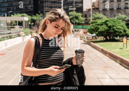 Une belle jeune femme d'affaires latine vérifiant les messages sur son téléphone portable avec un verre à la main pendant qu'elle marche au travail. Banque D'Images