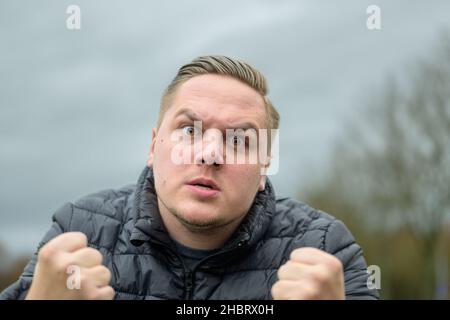 Un jeune homme furieux claque ses poings dans une rage et ronge ses dents alors qu'il menace le spectateur contre un ciel gris Banque D'Images