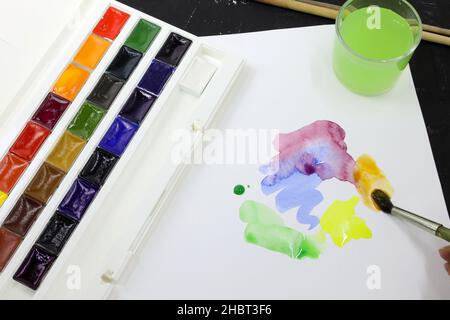 Peintures à l'aquarelle dans un contenant en plastique et peinture colorée sur une feuille.Peintures artistiques, un verre d'eau et une feuille de papier.Traits de pinceau o Banque D'Images
