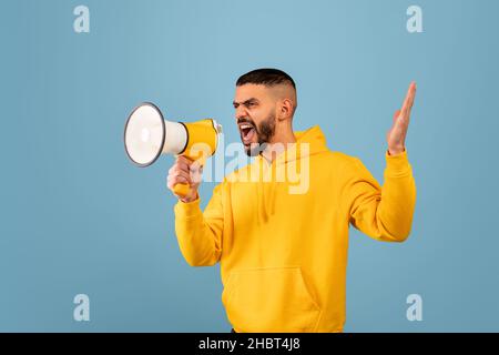 Portrait d'un jeune arabe agressif criant au mégaphone, debout sur fond bleu de studio Banque D'Images
