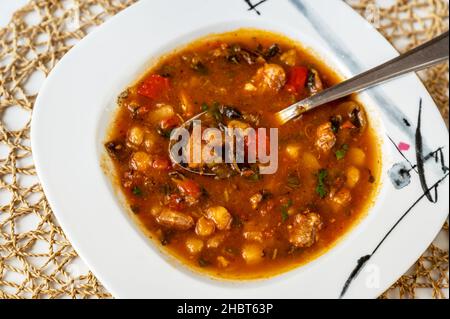 Soupe rouge rurale riche et épicée avec viande de porc, poivron rouge, tomate et champignons, légumes et haricots dans une assiette, cuillère, gros plan. Banque D'Images