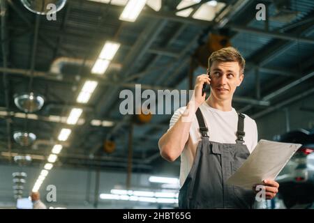 Vue en contre-plongée d'un jeune mécanicien professionnel, jeune homme, portant un uniforme tenant une planchette à pince et parlant sur un téléphone portable. Banque D'Images