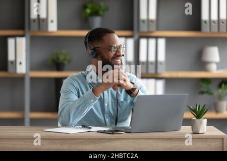 Un homme à barbe noire millénaire satisfait en lunettes et écouteurs travaille à l'ordinateur portable et regarde l'espace vide Banque D'Images