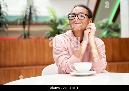 Noire africaine américaine femme avec vitiligo pigmentation problème de peau à l'intérieur vêtu de lunettes roses à capuchon sitiing table intérieur boisson thé sourire Heureux p Banque D'Images