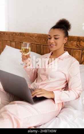 Jeune afro-américaine assise sur le lit avec un ordinateur portable et en dégustant du vin, en discutant vidéo avec des amis Banque D'Images