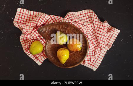 Vue de dessus, poires vertes et jaunes dans un bol en bois sculpté, nappe à carreaux rouges et panneau en marbre noir Banque D'Images