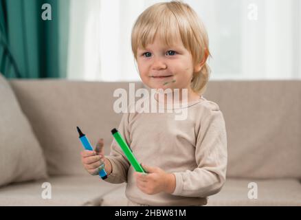 Une petite fille gaie en pyjama à la maison avec deux stylos feutre dans ses mains.Le visage et les vêtements sont tachés de peinture.Style de vie Banque D'Images