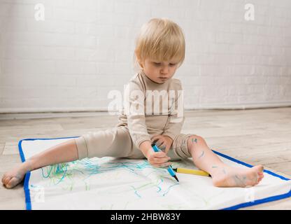 Une petite fille gaie en pyjama est assise sur le sol et de dessin avec des stylos-feutres sur un chiffon spécial.La face est tachée de peinture Banque D'Images