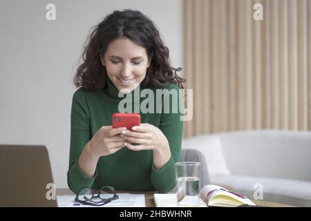 Jeune femme indépendante italienne avec des cheveux ondulés foncés tenant le téléphone cellulaire et regardant l'écran avec le sourire Banque D'Images