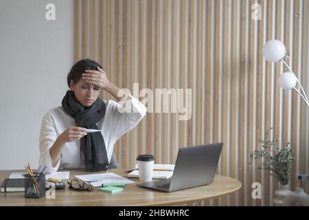 Femme fatiguée indépendante assise au bureau à la maison regardant malade au thermomètre numérique Banque D'Images