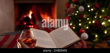 Détente de Noël à la maison.Homme avec un livre et un verre de brandy, foyer d'arrière-plan.Boire et lire, maison chaleureuse et confortable, vacances d'hiver. Banque D'Images