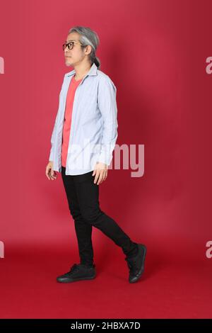 L'homme asiatique senior portant une chemise bleue avec de longs cheveux debout sur fond rouge. Banque D'Images