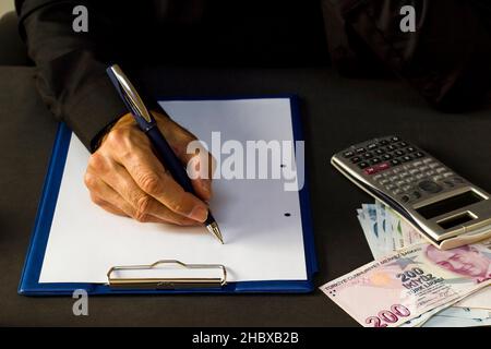 Homme main en chemise noire tenant stylo sur papier blanc vierge à la table noire avec quelques billets turcs et calculatrice. Banque D'Images