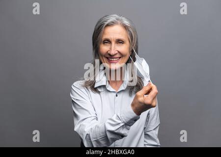 Heureuse femme âgée en tenue formelle qui a pris son masque médical isolé sur le gris.Portrait en gros plan d'une employée de bureau qui se protège contre les maladies virales.Recommande le concept des mesures Banque D'Images