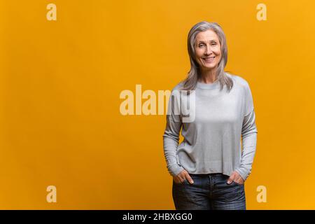 Une femme d'âge moyen garde les bras dans ses poches et pense à quelque chose tout en posant sur fond jaune.Femme âgée pensive avec cheveux gris Banque D'Images