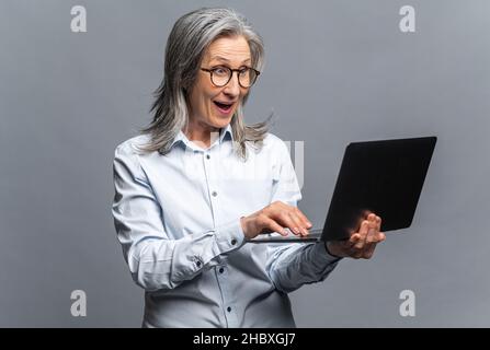 Surpris femme moderne mature utilisant un ordinateur portable isolé sur gris.Femme entrepreneur senior dactylographiant, femme d'âge moyen avec cheveux gris dans les e-mails de contrôle de port décontracté Banque D'Images