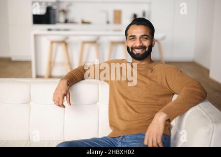 Un jeune Indien satisfait se reposant sur un canapé confortable dans un salon blanc confortable et moderne, regardant la caméra avec un sourire éclatant.Millennen hipster gars profiter de la vie sans stress sur le canapé à la maison Banque D'Images