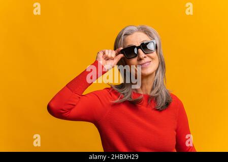 Taille haute vue portrait de la femme à la mode mature portant des lunettes de soleil posant à l'appareil photo sur le mur jaune.Photo Banque D'Images